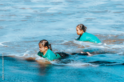 Two young children lie and play in shallow ocean waves near the shore, wearing matching green rash guards and enjoying a relaxed, playful summer moment at the beach.
