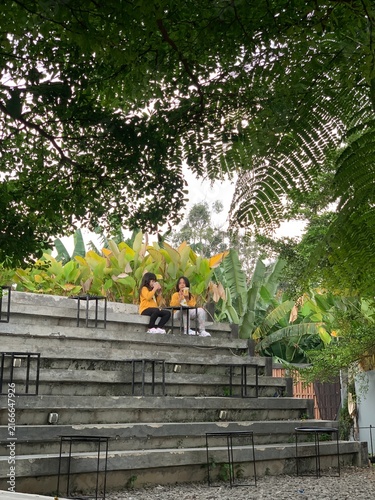 Two girls in matching yellow jackets sit on concrete terrace steps surrounded by lush tropical foliage, enjoying drinks, conversation and a relaxed outdoor moment in an urban garden setting.