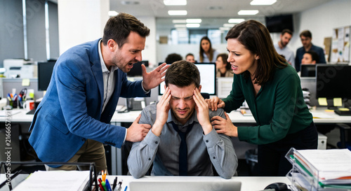 Stressed male employee holding his head while angry managers yell at him in a busy corporate office