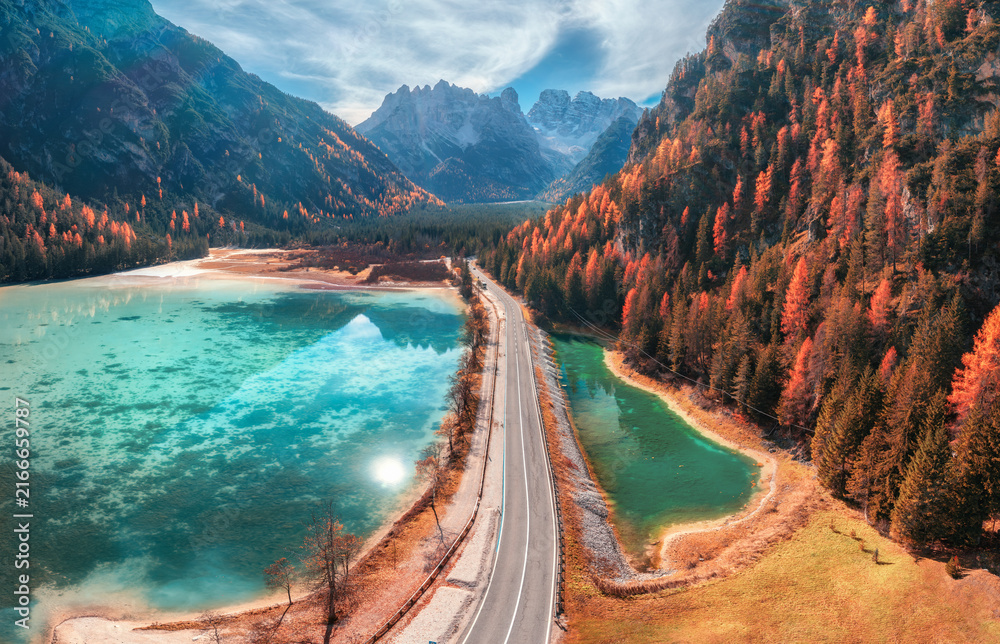 Fototapeta premium Aerial drone view of road along turquoise alpine lake in Dolomites, Italy. Coastal highway, mountain peaks, autumn larch trees forest slope, clear water, sky, clouds and reflection in fall. Top view