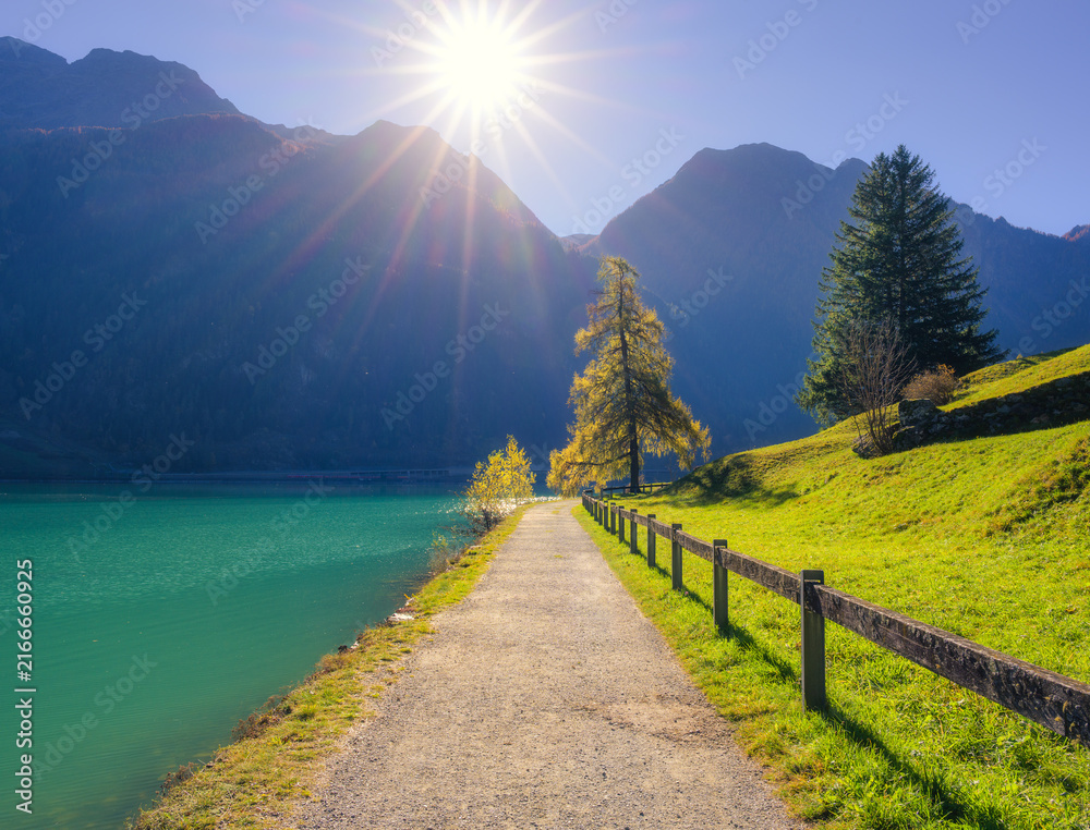 Fototapeta premium Hiking trail path along turquoise lake, trees and sun flare in autumn in Switzerland. Alpine landscape with sunbeams, larch tree, wooden fence, mountain peaks, green grass and blue water in fall
