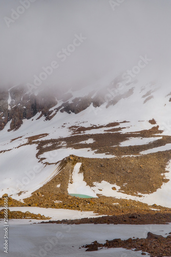 Foto Small turquoise glacial pond below melting snowfields and low clouds at Bogdanovich Glacier in Kazakhstan