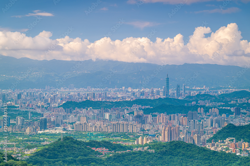 Fototapeta premium Scenic daytime aerial view of the Taipei city skyline, featuring modern skyscrapers, lush green hills, and distant mountains under a blue sky with fluffy white clouds.