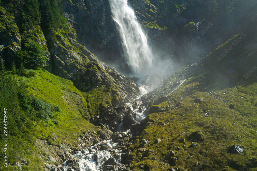 Fototapeta premium Waterfall rushes through a deep alpine gorge. Waterfall cascades over steep mountain rocks. Rushing water surrounds waterfall below slope. Rocky landscape frames waterfall in summer.