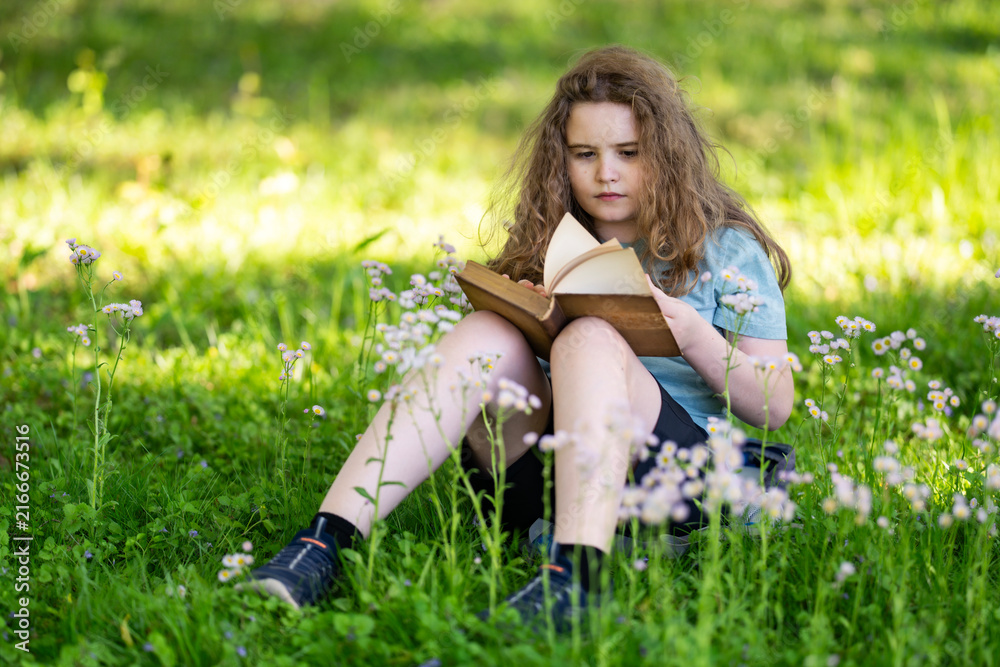 Fototapeta premium Child reading book in blurred bokeh. Young child enjoys reading book quietly. Educational portrait shows child reading book. Summer nature surrounds peaceful reading book.
