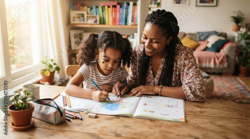 Caring mother and young daughter bonding while doing a school project at a sunny home table.