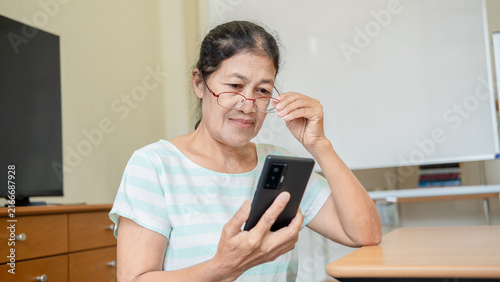 Canvas Print Elderly Asian woman using smartphone while adjusting glasses, Senior female using technology and with nearsighted or farsightedness, eye diseases