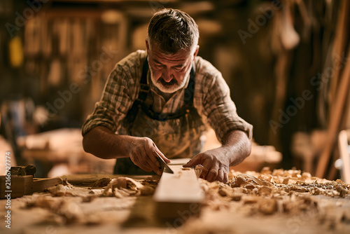 A senior Caucasian carpenter meticulously working on a wooden plank in a rustic workshop filled with tools and wood shavings.
