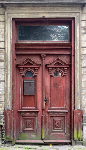 Ornate historic wooden double door with carved decorative panels, transom window, and a mounted mailbox, showing weathered red paint, aged timber, and traditional architectural craftsmanship.