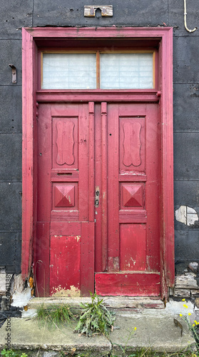 Weathered red wooden double door set within a dark aged façade, featuring peeling paint, geometric panels, a transom window, and signs of decay that reflect historic architectural character.