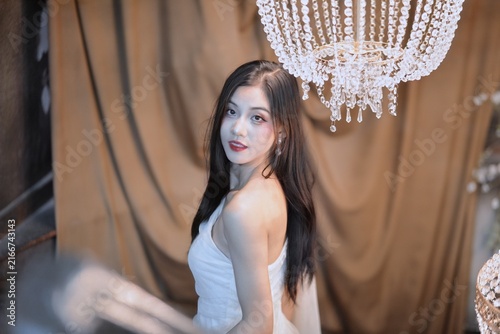 Elegant young Asian woman in white dress posing under crystal chandelier in vintage studio setting
