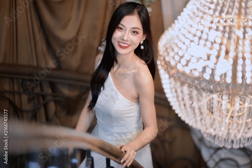 Elegant young Asian woman in white one-shoulder dress smiling on staircase beside crystal chandelier