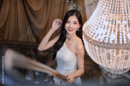 Elegant young Asian woman in white strapless dress smiling near luxury crystal chandelier in vintage interior