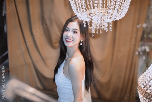 Smiling young Asian woman in elegant white dress posing under crystal chandelier at wedding venue