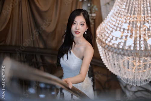 Elegant young Asian woman in white strapless gown standing on staircase beside luxurious crystal chandelier