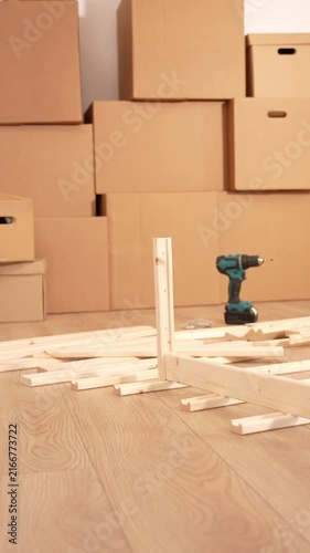Cardboard boxes stack in the background. Drill rests on wooden planks. The floor shows natural wood grain. The scene suggests moving or home projects. No people present