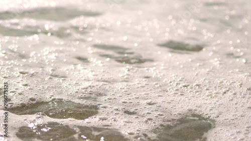 Close-up macro view of shimmering sea foam and tiny glistening bubbles washing over wet sand under bright daylight, capturing a tranquil mood.