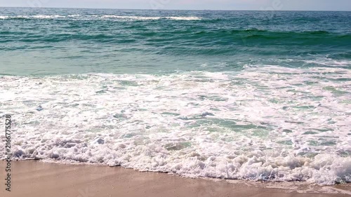 Gentle turquoise ocean waves roll onto a sunlit golden sand beach, creating white foam as they recede. Natural daylight illuminates the serene coastal landscape.