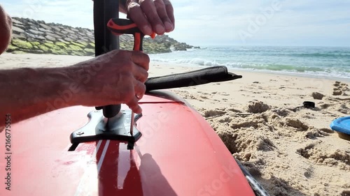 A surfer disassembles a hydrofoil mast from a red surfboard on a sandy beach. Bright sunlight illuminates the glossy surface and the ocean background.