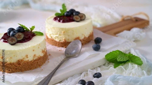 Two mini cheesecakes rest on parchment paper with vibrant berry sauce, fresh blueberries, and mint leaves, illuminated by soft natural lighting.