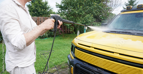 Washing yellow SUV car with high pressure washer gun. Man cleaning modern off-road vehicle with power washer in backyard garden, car detailing and maintenance at home.