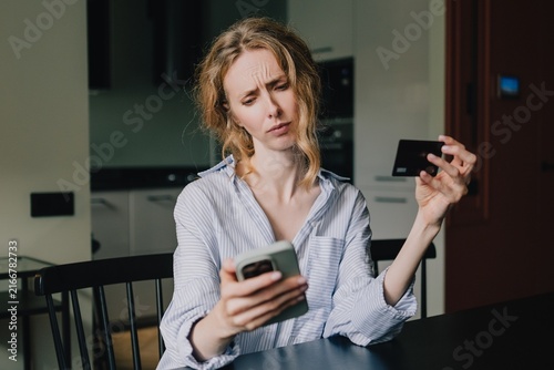 Woman looking concerned at smartphone while holding a bank card during an online payment problem at home