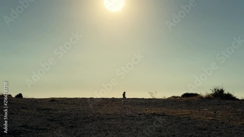 A cinematic video of a woman silhouette walking along a dry barren peninsula during warm summer evening sunset with a vast blue sky and serene coastal atmosphere.