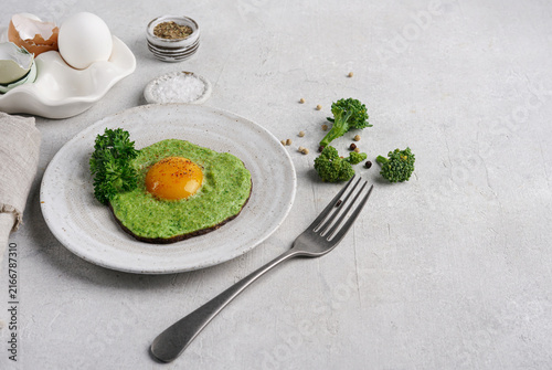 Healthy green fried egg with broccoli and spinach on white plate. Creative keto breakfast with egg yolk, herbs and spices on light textured background, copy space.