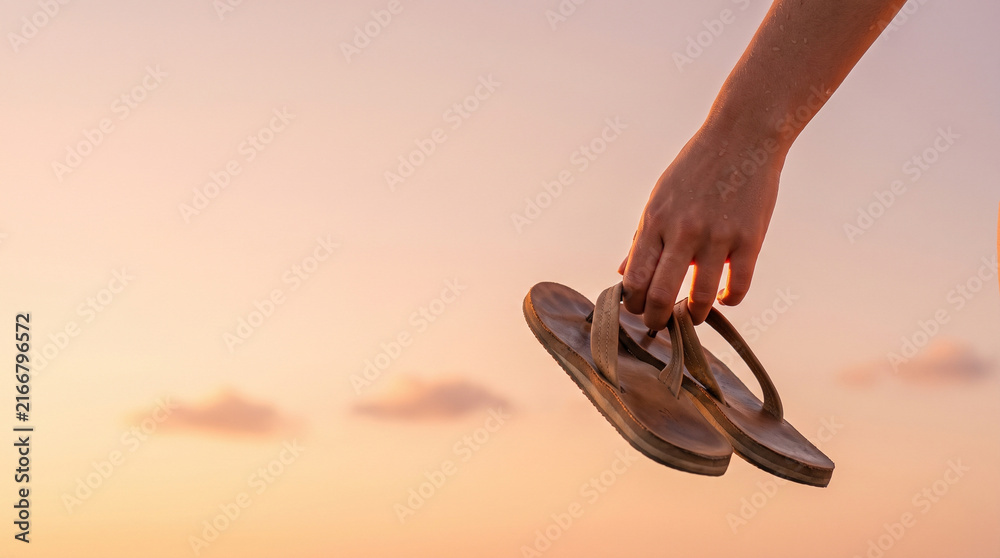 Naklejka premium Close Up of Hand Holding Leather Flip Flops Against Warm Sunset Sky, Concept of Summer Vacation, Freedom, Travel End, and Serene Tropical Beach Holiday with Copy Space