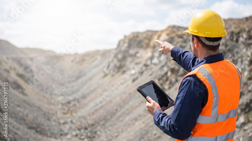 Rear View of Mining Engineer in Hard Hat and Safety Vest Holding Digital Tablet and Pointing at Open Pit Mine, Concept of Geological Survey, Industry Management with Copy Space