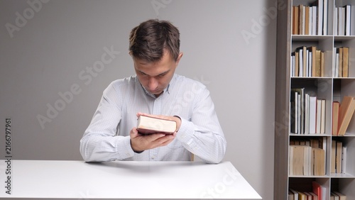 Obraz na płótnie young man sitting at a white desk in a library, gently holding and sniffing an o