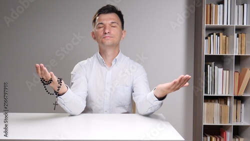 Фотография man with closed eyes holding rosary, palm raised in quiet prayer at a desk befor