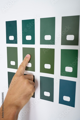 Hand points to one of several green paint color samples on white wall