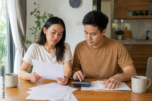 Young Southeast Asian couple reviewing monthly household bills with a calculator at their dining table