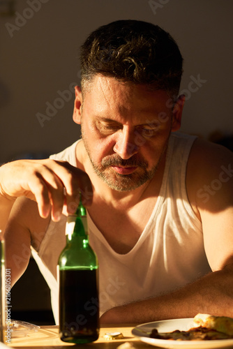 Photography Vertical image of middle aged Caucasian man sitting at table and holding beer bottle, looking down in despair