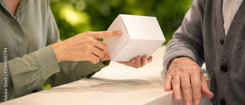 Close-up of customer service rep hands explaining product box to elderly couple — silver ring, wedding band, plant wall bokeh