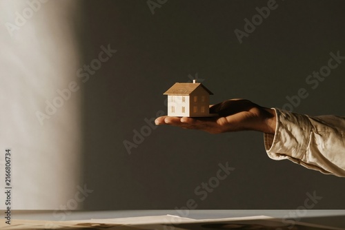 Small model house held in an open hand above a desk, featuring luminous lighting and soft textures for property advisory and mortgage consultation, with copy space