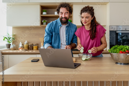 Obraz na płótnie Young couple cooking together in bright kitchen while following recipe on laptop