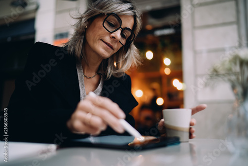 Close up of mature woman using stylus pen to sign digital document on tablet at outdoor cafe, focus on hand interaction with mobile screen, secure online transaction and banking concept