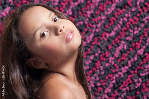 Close-up studio portrait of a young girl with long brown hair tilting her head with a soft expression against a pink textured background