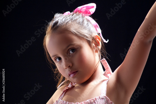 Close-up studio portrait of a young girl with pink ribbon in hair posing with raised arm on dark background