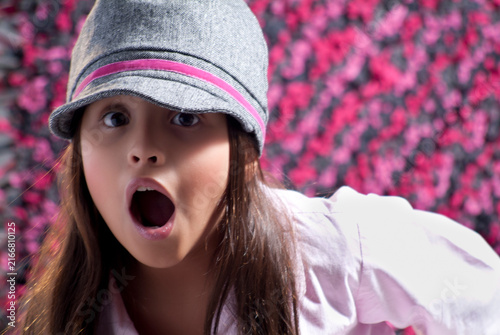 Surprised young girl wearing a grey cap with open mouth expression against a colorful pink textured background