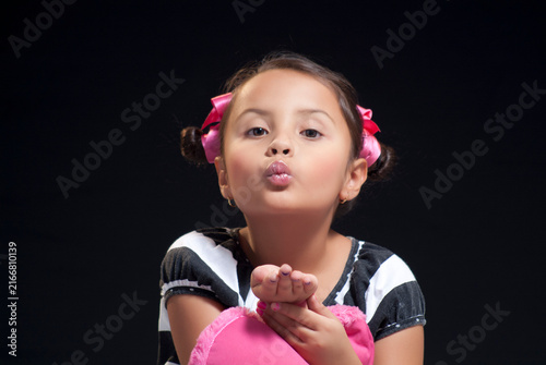 Adorable young girl with pink hair bows blowing a kiss toward the camera on dark background