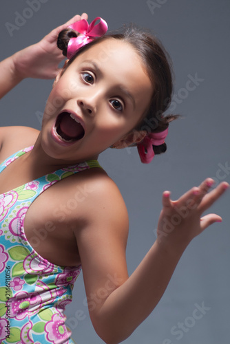 Excited young girl with pink hair bows making a playful surprised face gesture on grey background