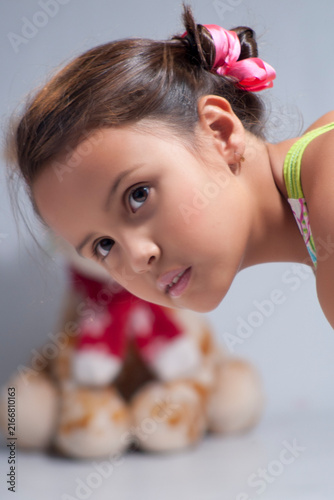 Curious young girl with pink hair ribbons leaning forward and looking closely with teddy bear blurred in background