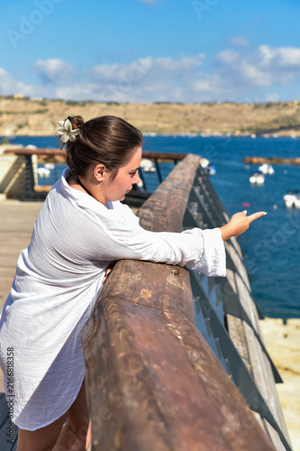 Fotografie A young woman leaning on a wooden railing points toward the sea in Malta