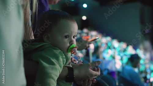 Baby in green hoodie held by adult watches stage performance in dark theater. Soft blue green ambient lighting. Background crowd blurred. Intimate family moment captured from side angle