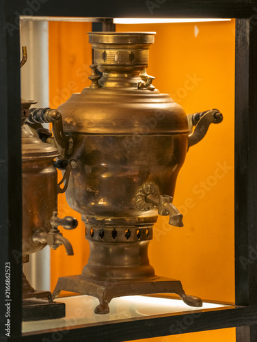 Foto Vintage brass metal Russian samovar water heater with a pouring spout standing on a shelf inside an illuminated dark wooden display cabinet