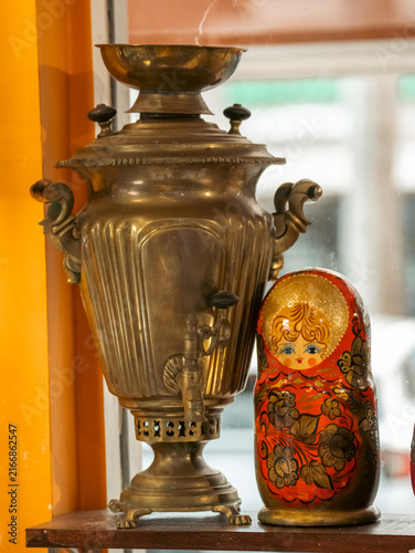 Photography Vintage brass metal Russian samovar water heater standing next to a traditional painted wooden matryoshka nesting doll on a wooden shelf