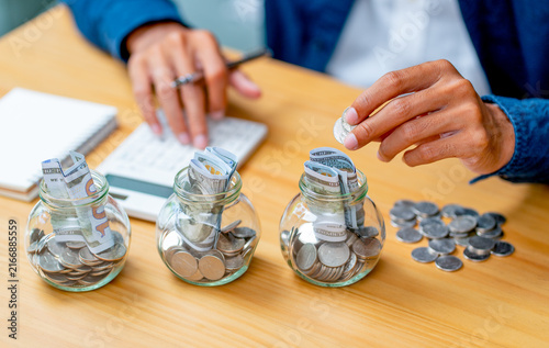 Person depositing coin into glass jar labeled 'PURPOSE' among three jars with rolled bills and coins on wooden desk